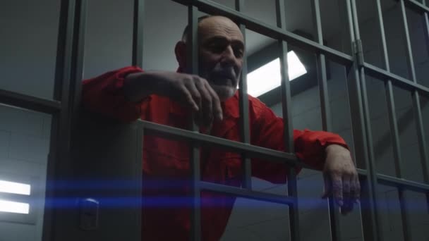 身穿橙色制服的老年囚犯手握金属棒 罪犯在狱中服刑 累坏了的囚犯在监狱 拘留中心或教养所的监牢中走着 — 图库视频影像