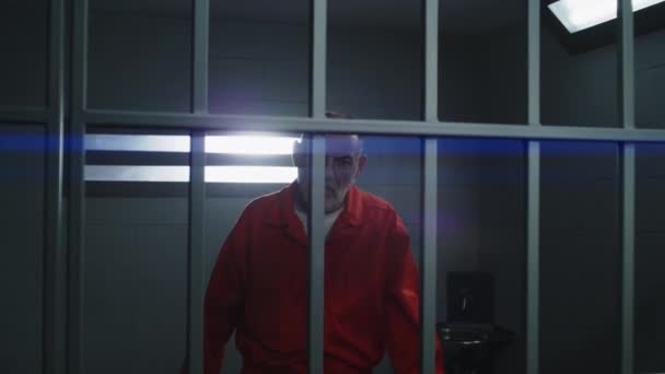オレンジの制服を着た高齢の犯罪者は 禁止された窓の近くに立って 刑務所のセルのバーにもたれ カメラを見ています 矯正施設の受刑者 囚人は懲役刑を言い渡される 肖像画 — ストック動画