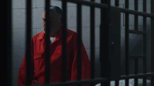 身穿橙色制服的老罪犯坐在监狱的床上 想着自由 犯人在监牢中服刑 在拘留中心或教养所的有罪囚犯 — 图库视频影像