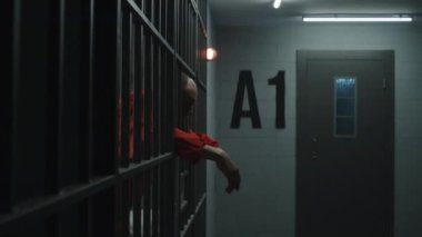 Turuncu üniformalı yaşlı mahkum ellerini uzatıyor, metal parmaklıklara yaslanıyor. Suçlu hapishane hücresinde hapis cezasına çarptırıldı. Hapisteki ya da ıslah evindeki üzgün mahkumlar.