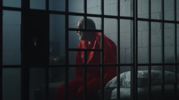 身穿橙色制服的老罪犯坐在监狱的床上 想着自由 抑郁的囚犯在监狱里服刑 拘留中心或教养所的有罪罪犯 — 图库视频影像
