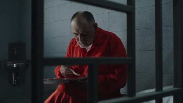 老年囚犯 身穿橙色制服的囚犯 坐在牢房里吃着食物 罪犯因犯罪而服刑 拘留中心或教养所 通过金属棒射击 — 图库视频影像