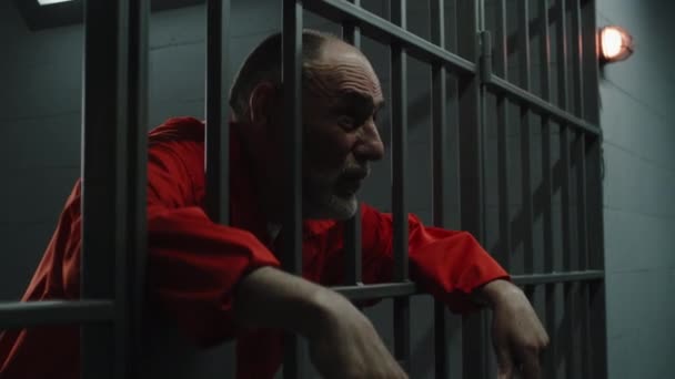 身穿橙色制服的老年囚犯伸出手指 靠在金属棒上 罪犯因犯罪在监牢服刑 累坏了的犯人站在监狱或拘留中心的铁窗后面 — 图库视频影像