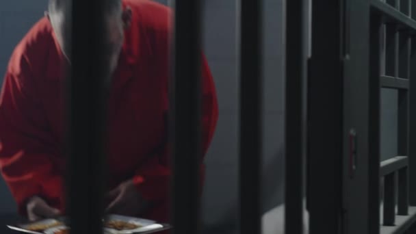 オレンジの制服を着た高齢の囚人が刑務所の独房に座っている 刑務官は金属棒を通して食物のトレイを与えます 受刑者は 夕食を食べ 刑務所又は矯正施設において懲役刑を言い渡される — ストック動画