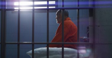 Duvardaki hapishane hücresinin numarası. Turuncu üniformalı yaşlı suçlu hapishane yatağında parmaklıklı pencereye bakıyor. Mahkum hapis cezasına çarptırıldı. Gözaltı merkezinde tutuklu var. Kapat..