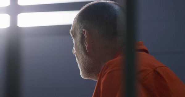 Elderly criminal in orange uniform sits on prison bed and dreams about freedom. Prisoner serves imprisonment term in jail cell. Gloomy inmate in detention center or correctional facility.