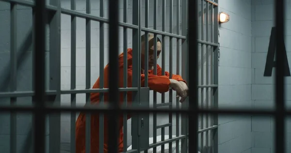 オレンジの制服を着た高齢の囚人は 金属の棒にもたれて指を伸ばします 刑事は刑務所の独房で犯罪の刑期を務める 被収容者は刑務所や拘置所のバーの後ろに立っています — ストック写真