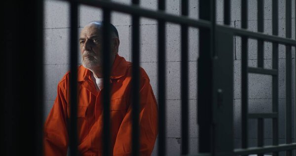 Elderly criminal in orange uniform sits on prison bed and thinks about freedom. Prisoner serves imprisonment term in jail cell. Guilty inmate in detention center or correctional facility.
