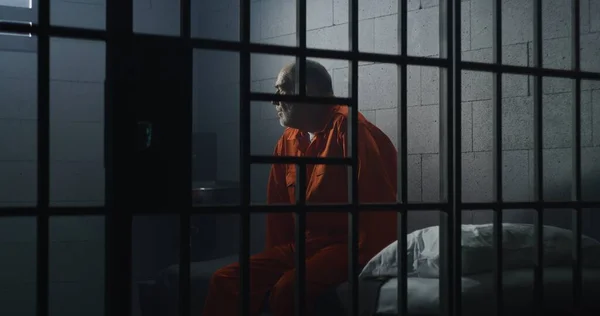 身穿橙色制服的老罪犯坐在监狱床上 与狱友交谈 沮丧的囚犯在监狱里服刑 拘留中心或教养所的有罪囚犯 — 图库照片