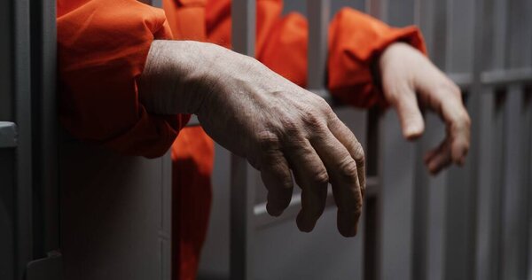 Hands close up of elderly prisoner in orange uniform holding metal bars, standing in prison cell. Criminal serves imprisonment term for crime. Inmate in jail or correctional facility. Justice system.