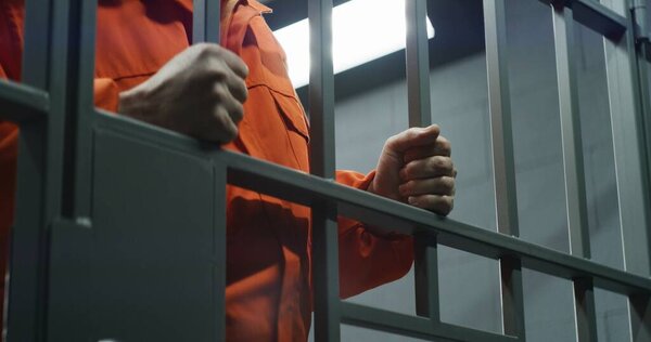 Close up of prisoner in orange uniform holding metal bars, standing in prison cell. Guilty criminal or killer serves imprisonment term for crime. Inmate in jail or detention center. Justice system.