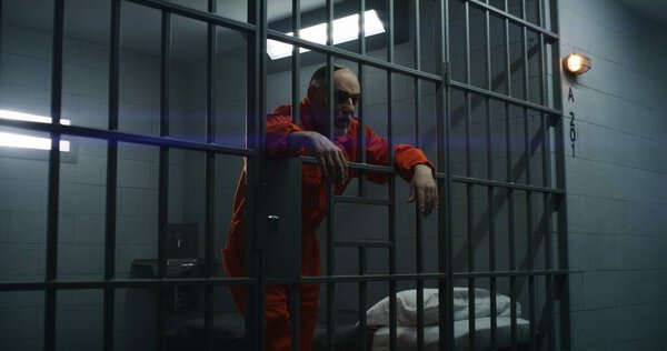 Elderly prisoner in orange uniform leans hands on metal bars. Criminal serves term of imprisonment in prison cell. Gloomy inmate stands behind bars in jail, detention center or correctional facility.