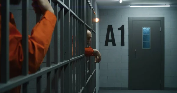 身穿橙色制服的老年囚犯伸出手指 靠在金属棒上 罪犯因犯罪在监牢服刑 累坏了的犯人站在监狱或拘留中心的铁窗后面 — 图库照片