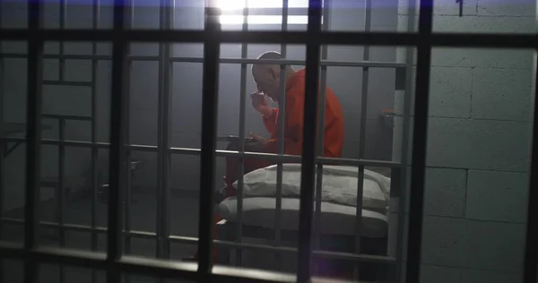 身穿橙色制服的老年囚犯坐在监牢里 吃着食物 罪犯因犯罪而服刑 监狱或教养所的条件 通过金属棒从牢房开枪射击 — 图库照片
