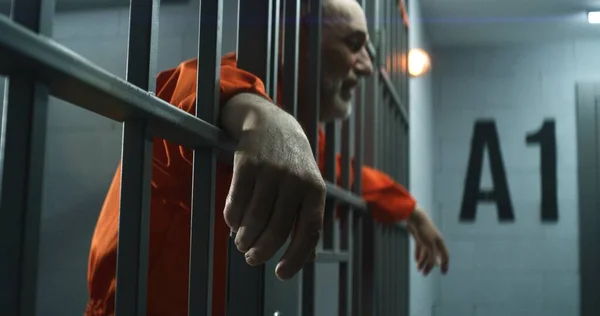 オレンジの制服を着た高齢の囚人は彼の手を伸ばし 金属棒にもたれる 刑事は刑務所の独房で刑期を務める 仮釈放者は 刑務所又は矯正施設のバーの後ろに立つ — ストック写真
