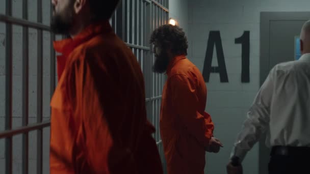 オレンジの制服を着た2人の囚人が刑務所の独房の前の金属の棒に面して立っている 警察のバトンウォークを持つ刑務所の役員は 刑務所で犯罪者を監視します 収容所又は矯正施設 — ストック動画