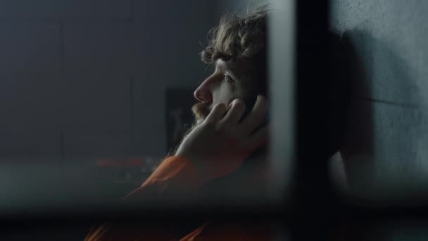 身穿橙色制服的囚犯躺在床上 在牢房里打电话 罪犯在狱中服刑 在拘留中心 教养所的囚犯 通过金属棒观看 靠近点 — 图库视频影像
