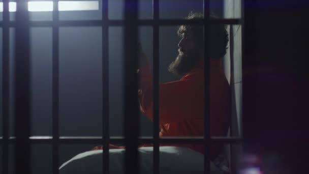 身穿橙色制服的囚犯坐在床上 在装有铁窗的牢房里通过电话交谈 有罪的罪犯在狱中服刑 拘留中心或教养所的囚犯 — 图库视频影像