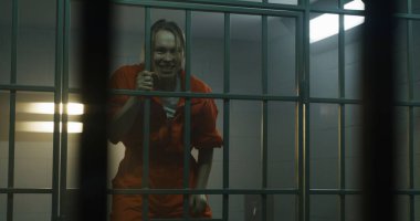 Turuncu üniformalı deli kadın mahkûm komşu mahkuma bağırıyor, hapishane hücresindeki parmaklıklara atlıyor. Kadın suçlu, hapiste suçtan hapis yatıyor. Gözaltı merkezi. Metal çubuklardan bak.