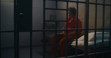 Turuncu üniformalı mahkum, yemek servisinden hapishane hücresindeki kadın suçluya yemek veriyor. Kadın suçlu yatakta oturuyor, hapis ya da gözaltı merkezinde hapis yatıyor..