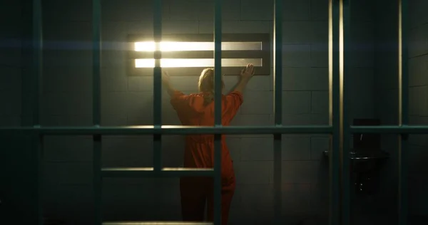 オレンジの制服を着た女性が歩き 金属の棒の後ろに立って 刑務所の独房の禁止された窓を見ている 女性は懲役刑を言い渡される 拘置所に収容されている — ストック写真