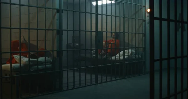 Jedna Więźniarka Pomarańczowym Mundurze Stoi Metalowymi Kratami Druga Siedzi Łóżku — Zdjęcie stockowe