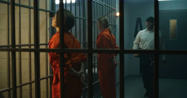 2人の女性囚人 オレンジの制服の受刑者は刑務所のセルの前の金属棒に直面して立つ 刑務所の役人が歩いて刑務所の女性犯罪者を見てる 収容所又は矯正施設 — ストック写真