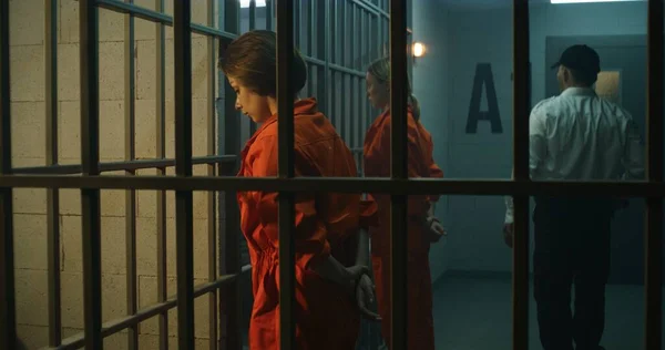 2人の女性囚人 オレンジの制服の受刑者は刑務所のセルの前の金属棒に直面して立つ 刑務所の役人が歩いて刑務所の女性犯罪者を見てる 収容所又は矯正施設 — ストック写真