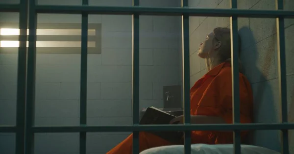 オレンジの制服を着た女性は金属棒の後ろのベッドの上に座り 刑務所の独房で聖書を読み取り 禁止された窓を見ています 女子犯罪者は 刑務所又は矯正施設において刑を言い渡される — ストック写真