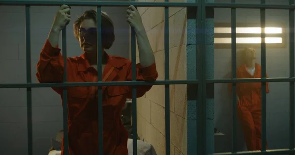 オレンジの制服を着た囚人の一人が金属棒の後ろに立ち もう一人は独房のベッドの上に座っている 女性は刑務所で犯罪の懲役刑を言い渡される 拘置所の被収容者 — ストック写真