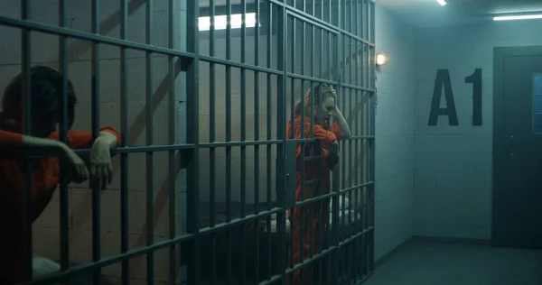 Jedna Więźniarka Pomarańczowym Mundurze Stoi Metalowymi Kratami Druga Siedzi Łóżku — Zdjęcie stockowe