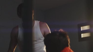 Angry prisoner talks aggressively to cellmate, threatens him in prison cell. Inmates serve imprisonment terms for crimes in jail. Detention center or correctional facility. View through metal bars.
