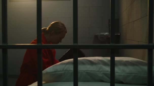 ベッドの近くのオレンジの制服の膝の女性の囚人は 聖書を保持する刑務所の独房で神に祈ります 女子犯罪者は 刑務所 拘置所又は矯正施設において懲役刑を言い渡される — ストック動画