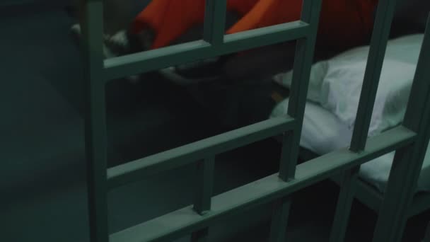 オレンジ色の制服を着た男性は刑務所に座っている ジャイラーは金属棒を通して聖書を与えます インメイトは刑務所や矯正施設で刑務所に収監される 神の概念に対する信仰 クローズアップ — ストック動画