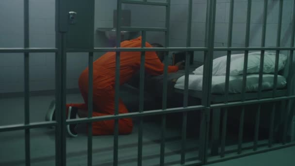 ベッドの近くでオレンジ色の制服を着たアフリカ系アメリカ人の囚人は 聖書を保持する刑務所で神に祈ります ジャイラーが歩き回り 刑務所や矯正施設で犯罪者を見ている 神への信仰 — ストック動画