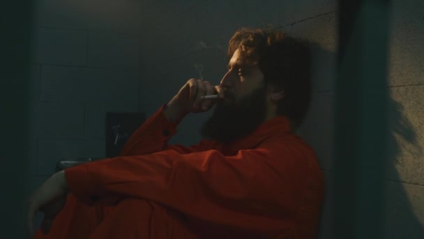 身穿橙色制服的沮丧的男性囚犯坐在床上 在监狱里抽烟 犯人因犯罪在监狱服刑 阳光透过有栅栏的窗户照耀着 拘留中心 — 图库视频影像