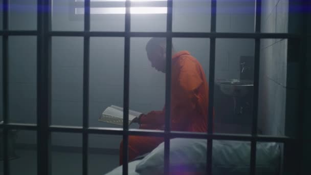 Prisionero Afroamericano Con Uniforme Naranja Sienta Cama Lee Biblia Celda — Vídeos de Stock