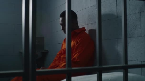オレンジ色の制服を着た有罪のアフリカ系アメリカ人は刑務所のベッドに座り 自由について考えている 矯正施設で抑圧された犯罪者 刑務所刑務所の刑務所に収監されている ドリー ショット — ストック動画