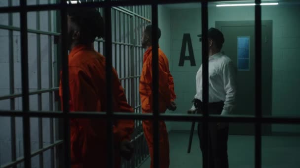 两名身穿橙色制服的囚犯面对着铁棍站在牢房前 拿着警棍的狱卒跟在罪犯后面 与他们交谈 杀人犯在拘留中心服刑 — 图库视频影像