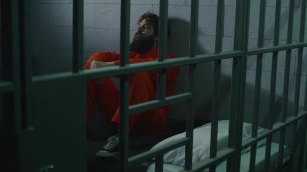 オレンジ色の制服を着た男性は 刑務所のベッドに座っている ジャイラーは金属棒を通して聖書を与えます 刑務所では刑務所で有罪判決が下される 拘禁センターまたは矯正施設 — ストック動画