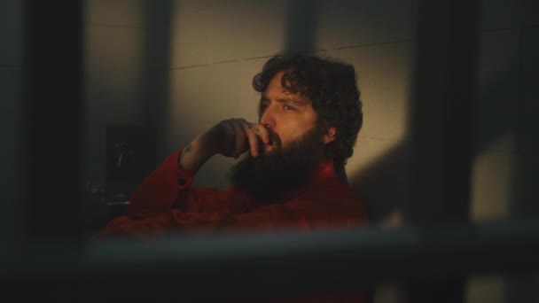 身穿橙色制服的沮丧囚犯坐在床上 在监狱里抽烟 犯人因犯罪在监狱服刑 阳光透过有栅栏的窗户照耀着 惩教设施 — 图库视频影像