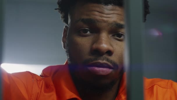 Afroamerykanin Pomarańczowym Mundurze Stoi Kratkami Więziennymi Patrzy Kamerę Więzień Odsiaduje — Wideo stockowe