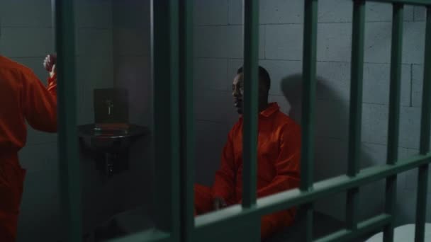 オレンジ色の制服を着た2人のアフリカ系アメリカ人の囚人が話す 一人の男がベッドに座り もう一人は刑務所の中を歩いている 収容所または矯正施設における犯罪の刑期を定める — ストック動画