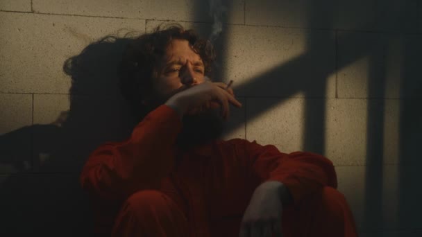 身穿橙色制服的沮丧囚犯坐在床上 在监狱里抽烟 犯人因犯罪在监狱服刑 阳光透过有栅栏的窗户照耀着 惩教设施 — 图库视频影像