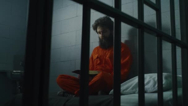 身穿橙色制服的男性囚犯坐在床上 看着牢笼中的窗玻璃 犯人因犯罪在狱中服刑 惩教设施 对上帝的信仰概念 — 图库视频影像