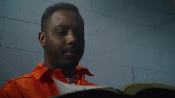 African American Prisoner Orange Uniform Sits Prison Cell Reads Book — Stock Video