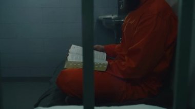 Turuncu üniformalı bir erkek mahkum yatakta oturur, hücrede İncil okur. Suçlu, mahkum hapiste suçtan hapis cezasına çarptırıldı. Gözaltı merkezi ya da ıslah evi. Tanrı kavramına güven.