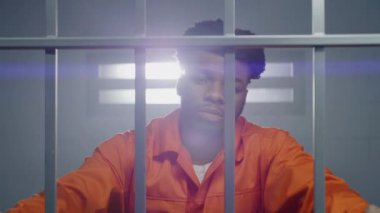 Turuncu üniformalı Afro-Amerikalı bir adam hapishane hücresinde kameraya bakar ve boynu yoğurur. Mahkum hapiste hapis yatar. Islah evinde ya da gözaltı merkezinde bir suçlu. Dikey görünüm.