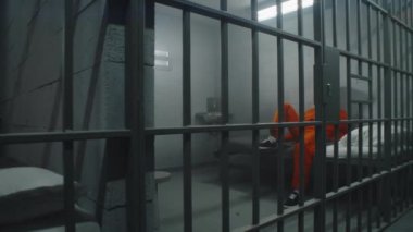 İki komşu mahkum hapishane hücrelerinde hapis yatıyor. Turuncu üniformalı Afro-Amerikan suçlu hapishane yatağında parmaklıklı pencereye bakıyor. Gözaltı merkezindeki kasvetli mahkumlar. Dolly vurdu..
