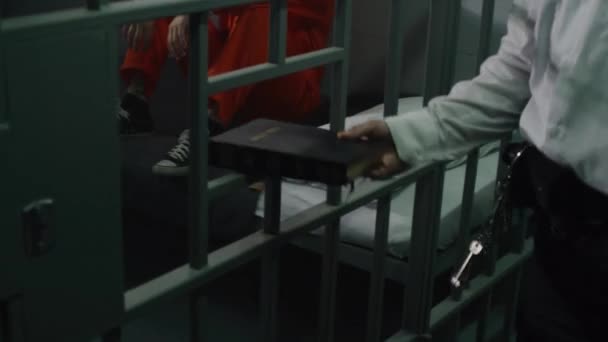 监狱官发给身穿橙色制服的男性囚犯圣经 罪犯坐在床上的牢房 罪犯因在狱中犯罪而被判处监禁 拘留中心 对上帝的信仰概念 — 图库视频影像